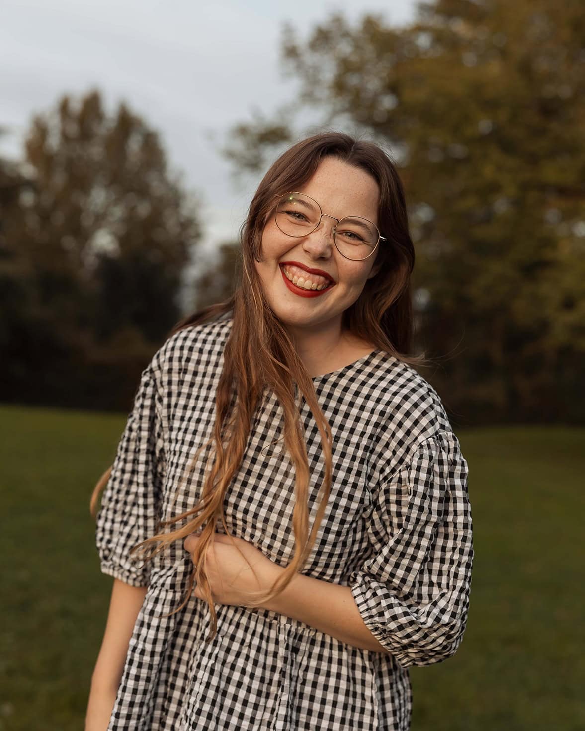 Helene smiler bredt i et utendørs landskap, iført rutete kjole og briller, omgitt av høstfargede træ.