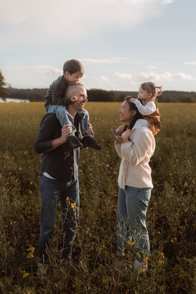 Familie med to voksne og to barn i blomstereng under solskinn.