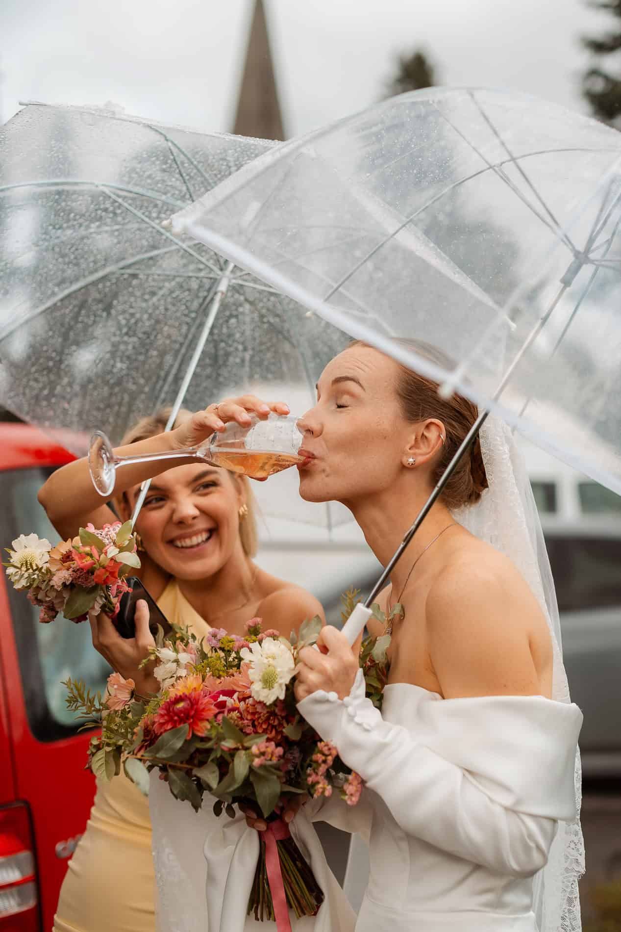 Brud og venninne nyter drikke under paraplyer på bryllupsdag.