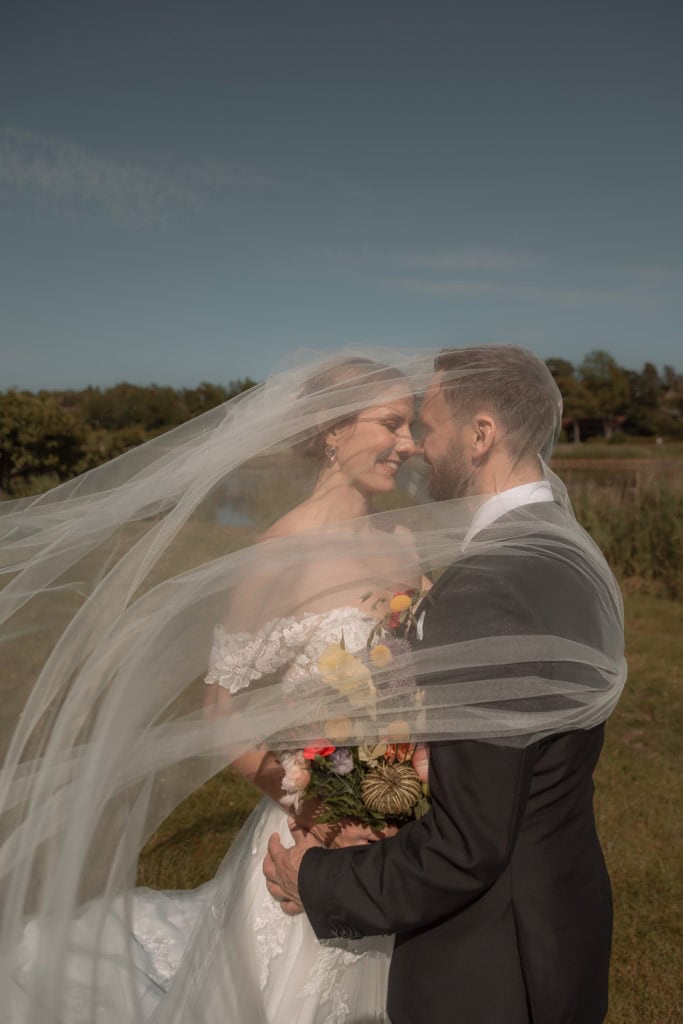 Bryllupsfoto av et lykkelig brudepar som deler et øyeblikk under en åpen himmel, med brudens slør so.