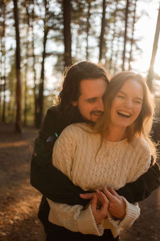 Voksenpar i skog, smilende og omfavnet, nyter naturen og hverandre i solskinnet.