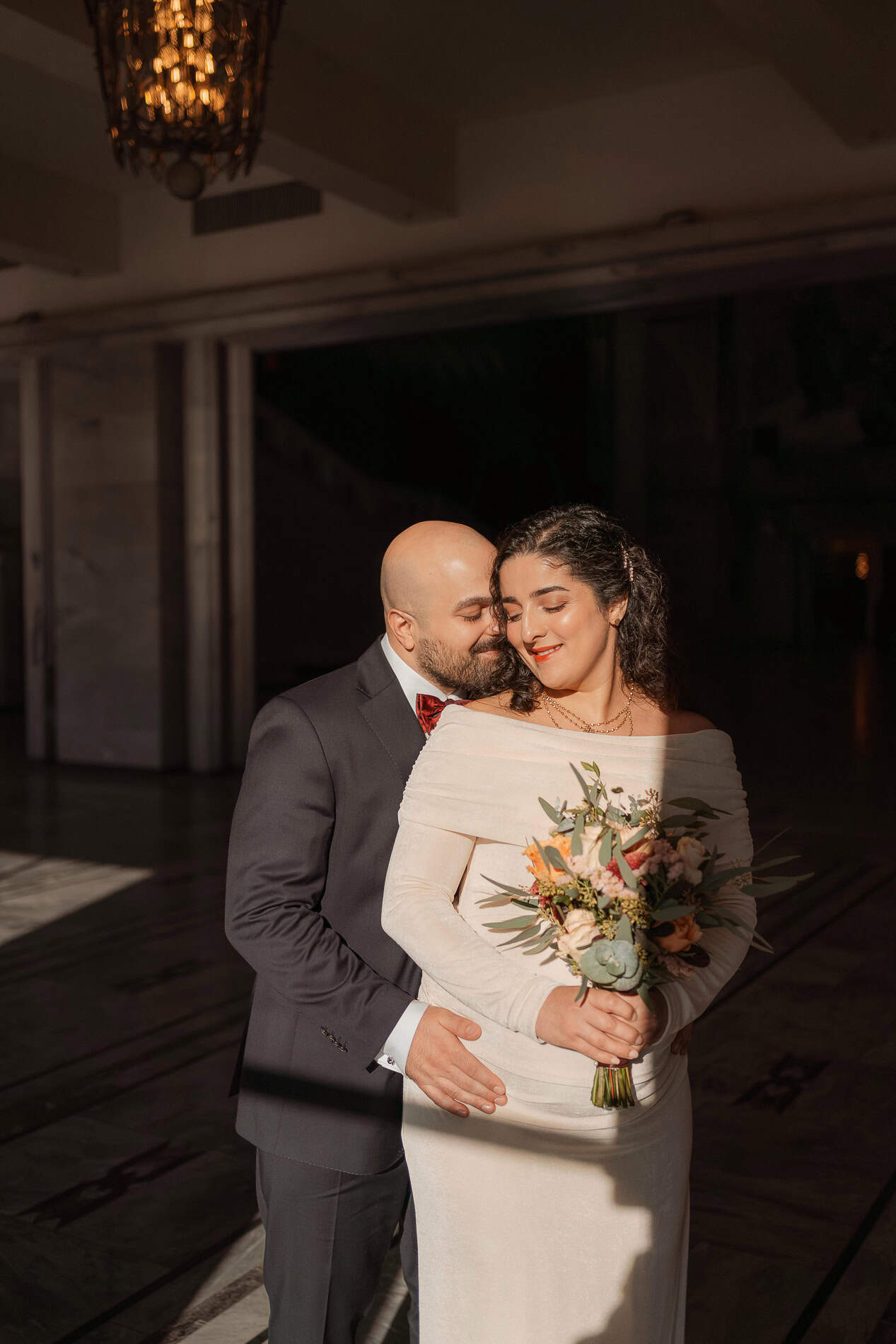 Par i bryllupsklær som kysser og smiler, med blomsterbukett, i et elegant innendørs bryllupsrom.