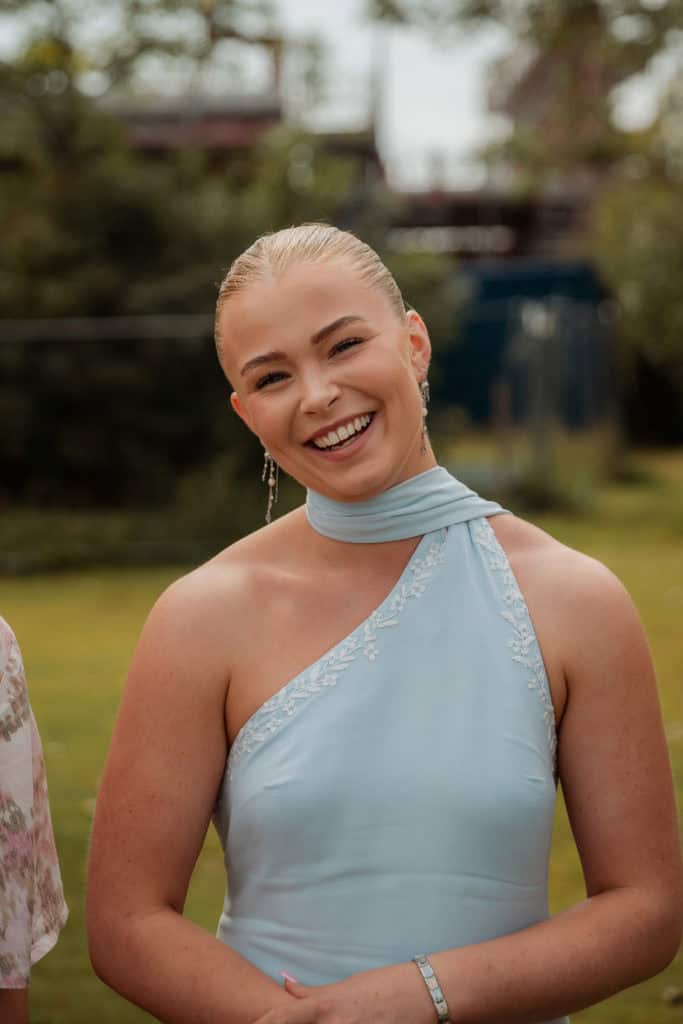 Brudepike smiler lykkelig i lyseblå kjole under bryllupet, omgitt av vakker natur og feststemning.