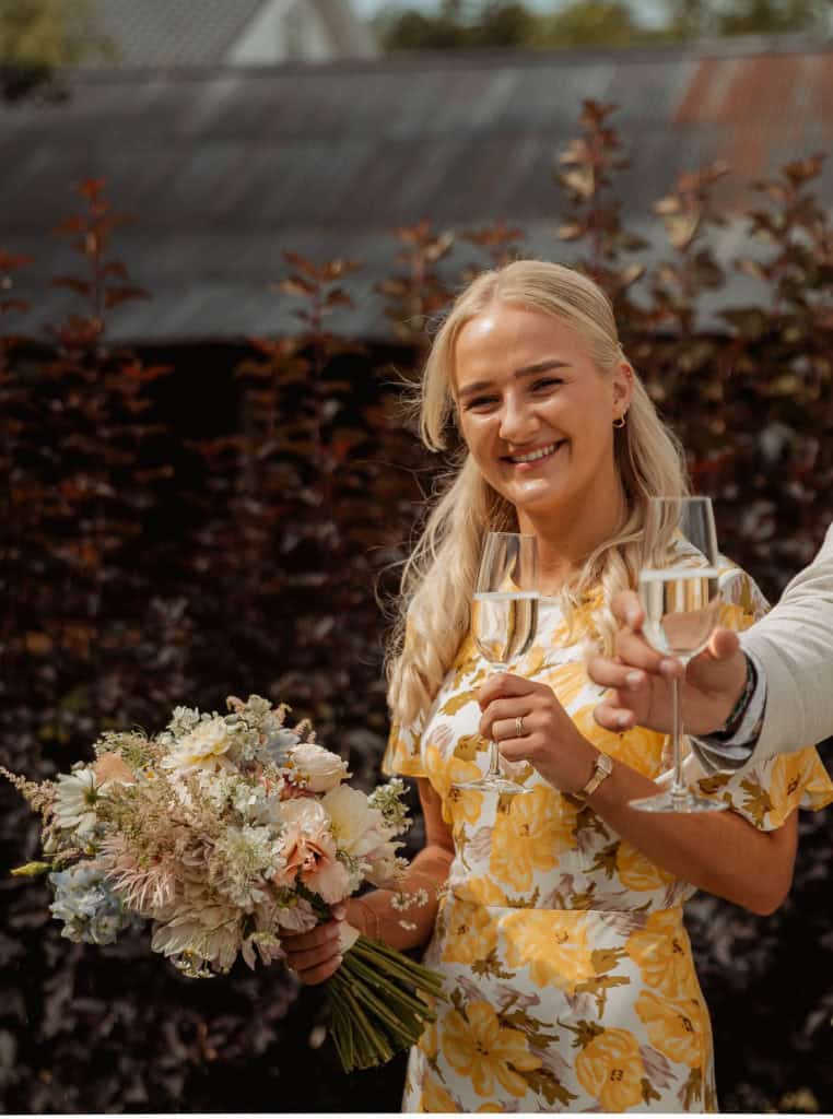 Bryllupsstemning med smilende brud som holder blomsterbukett og skåler med champagne under bryllupet.