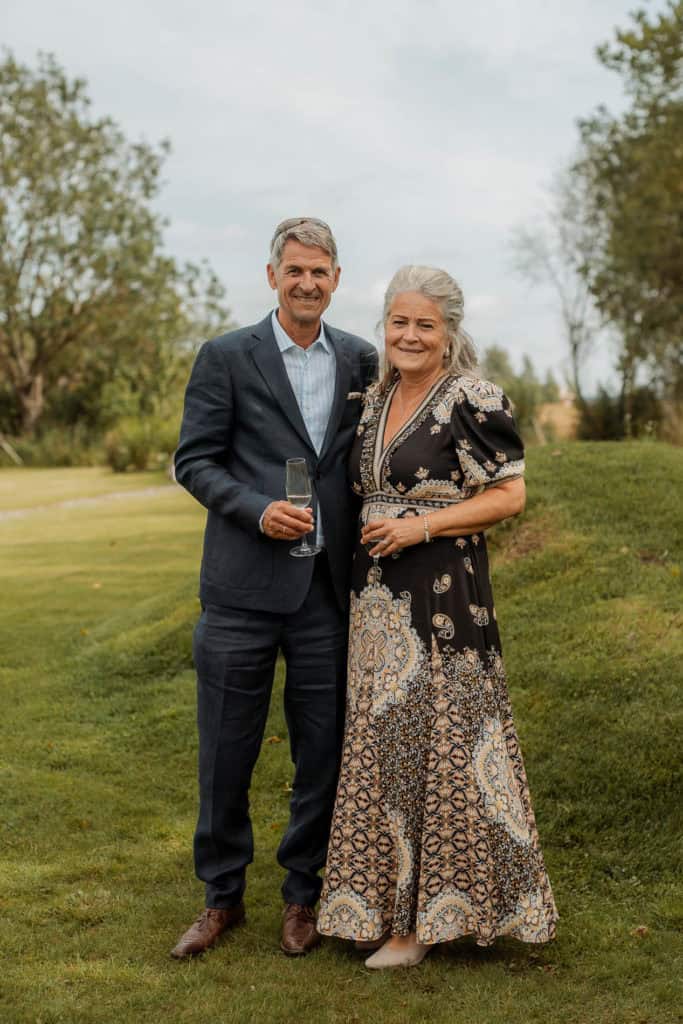 To personer i bryllupskledning smiler og holder glass, ute i naturen under bryllupsfeiring.