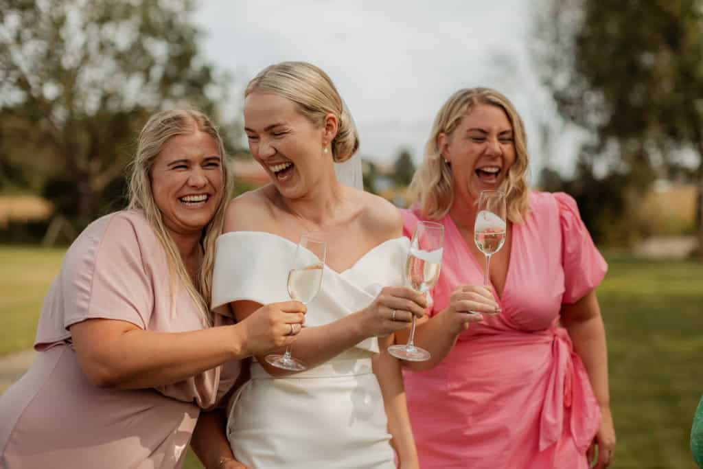 Tre kvinner i bryllupsklær smiler og skåler under bryllupet, glede og vennskap.