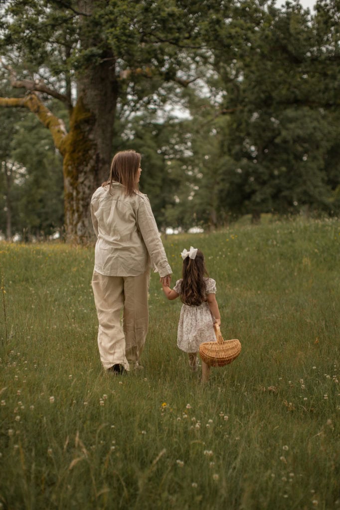 Voksen og barn går hånd i hånd i en grønn eng med trær i bakgrunnen.