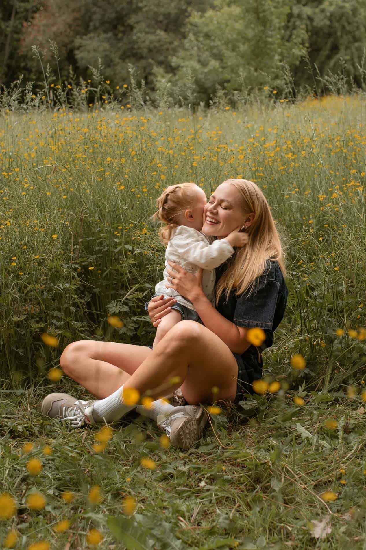 Voksen kvinne og liten jente koser i blomstereng med gult blomsterteppe.