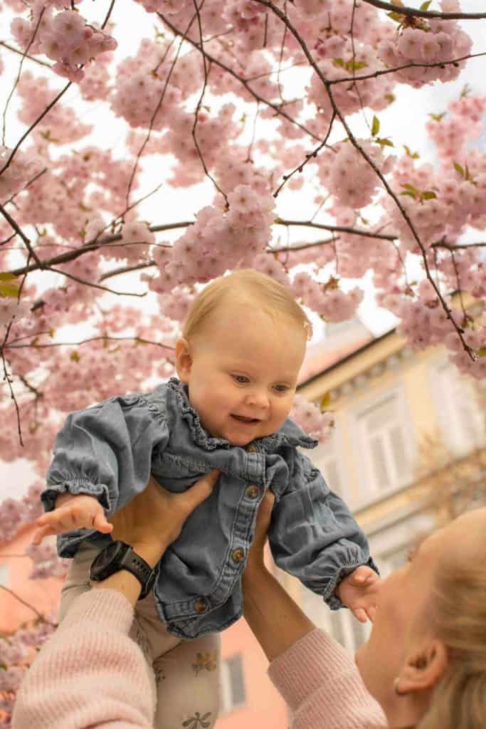 Barneløft under blomstrende kirsebærtrær, smilende baby og omsorgsfull voksen i vårlige omgivelser.
