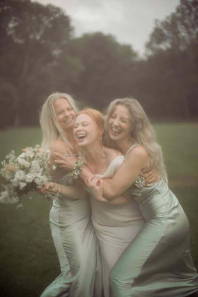 Tre kvinner i blomsterkjoler omfavner hverandre i en park, smilende og glade.