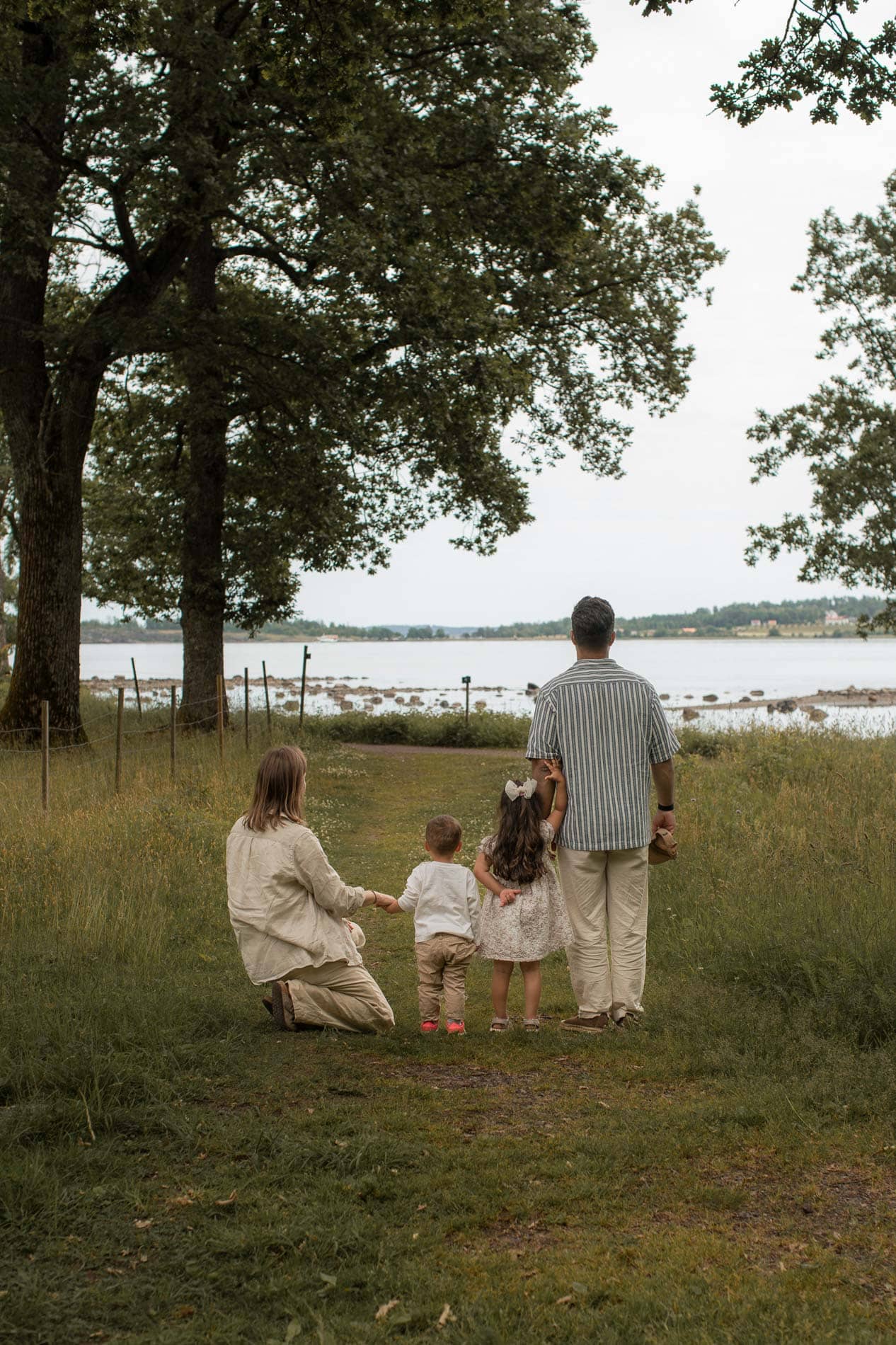 Familie som går langs sjøen med barn, omgitt av grønne trær og åpen himmel.