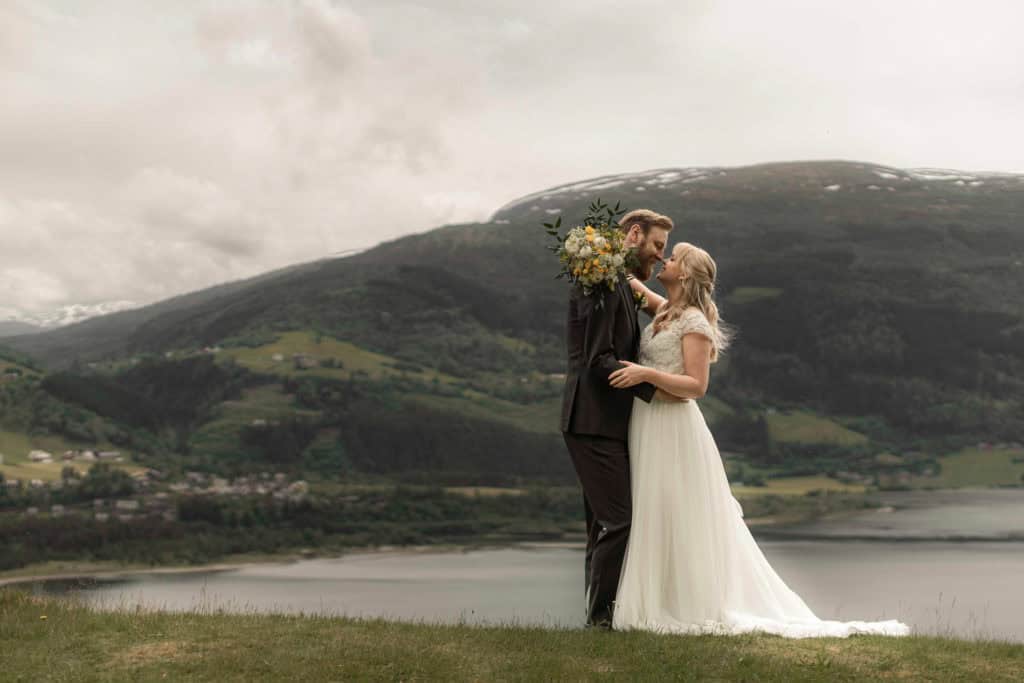 Romantisk bryllupsbilde av et par som kysser ved innsjøen med fjell i bakgrunnen.