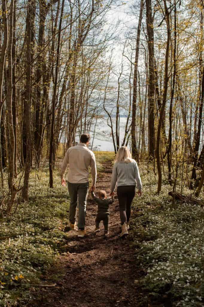 Familie går tur i skogen med barn og hund, omgitt av vårblomster og trær.
